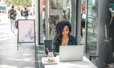 ethnic young woman using laptop while having tasty beverage in modern street cafe